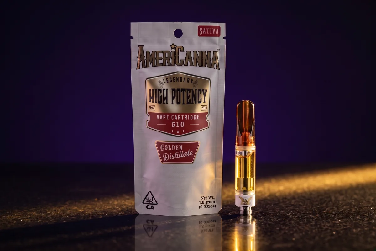 A silver AmeriCanna Sativa 510 vape cartridge pouch, 1.0 gram of golden distillate, with one cartridge of golden oil beside it, on dark stone.