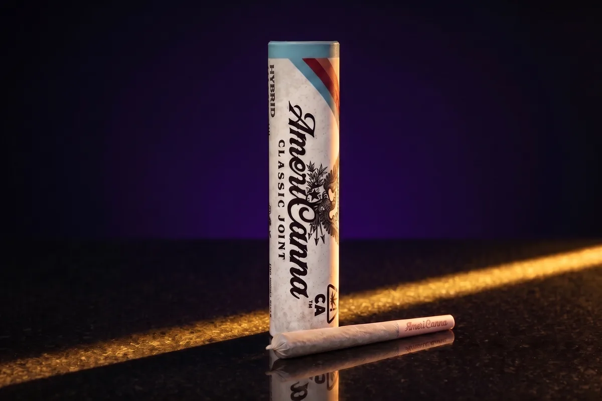 An AmeriCanna Classic Joint pre-roll tube, off-white with a teal top band, with one unlit pre-roll in front, on black stone in gold light.