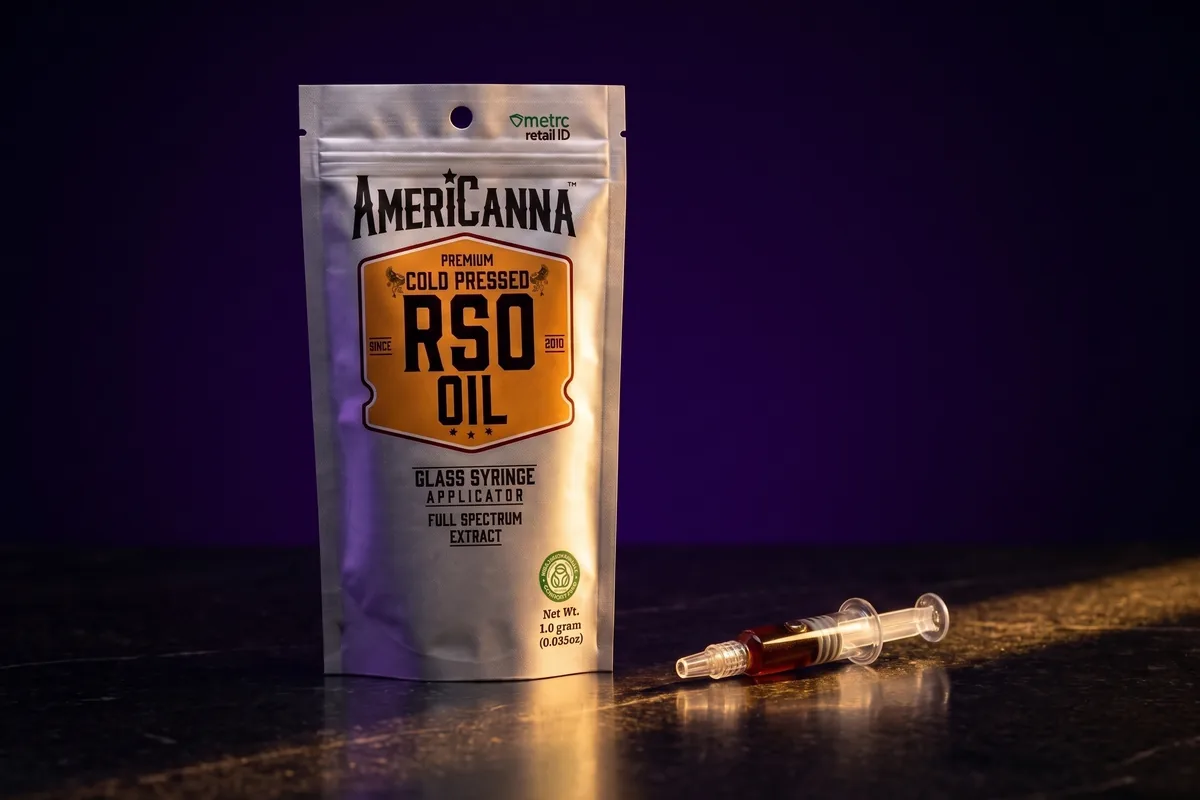 A silver AmeriCanna Premium Cold Pressed RSO Oil pouch, 1.0 gram, with a glass syringe of dark oil beside it, on black stone in gold light.