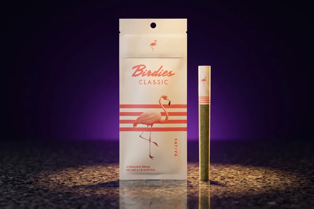 A cream Birdies Classic 3-pack pouch, Sativa, with coral stripes and a pink flamingo, and one pre-roll standing beside it on dark granite.