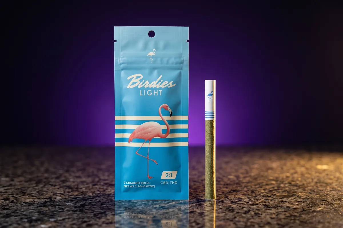 A sky-blue Birdies Light 3-pack pouch, 2:1 CBD to THC, with a pink flamingo, and one pre-roll with blue rings beside it on dark granite.