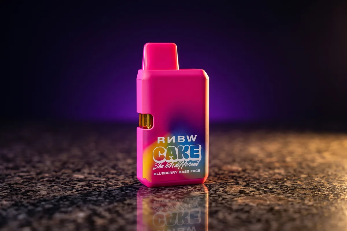 One hot-pink CAKE RNBW 1.25g all-in-one vape in Blueberry Bass Face, with a rainbow wash on its front, standing on dark granite in gold light.