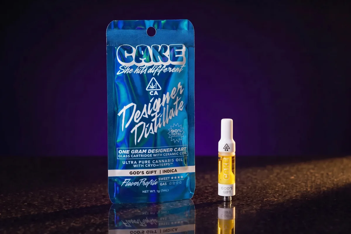 A blue holographic CAKE Designer Distillate cart pouch in God's Gift, Indica, with one glass cartridge of golden oil beside it on dark stone.