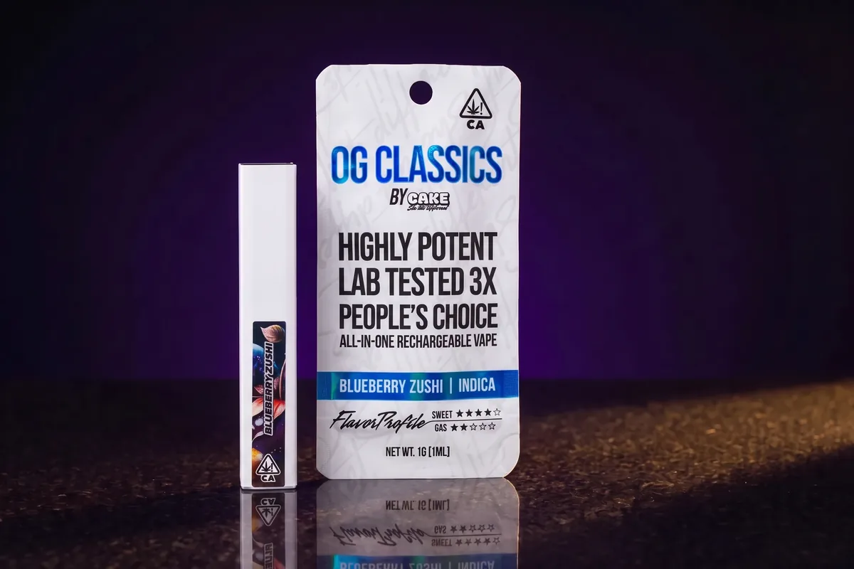 A white CAKE OG Classics pouch in Blueberry Zushi, Indica, a 1g all-in-one rechargeable vape, with the slim white pen beside it on dark stone.