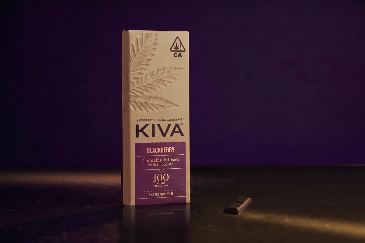 A Kiva Blackberry dark chocolate bar box, tan kraft with a plum label, and one segment of dark chocolate in front, on dark stone in gold light.