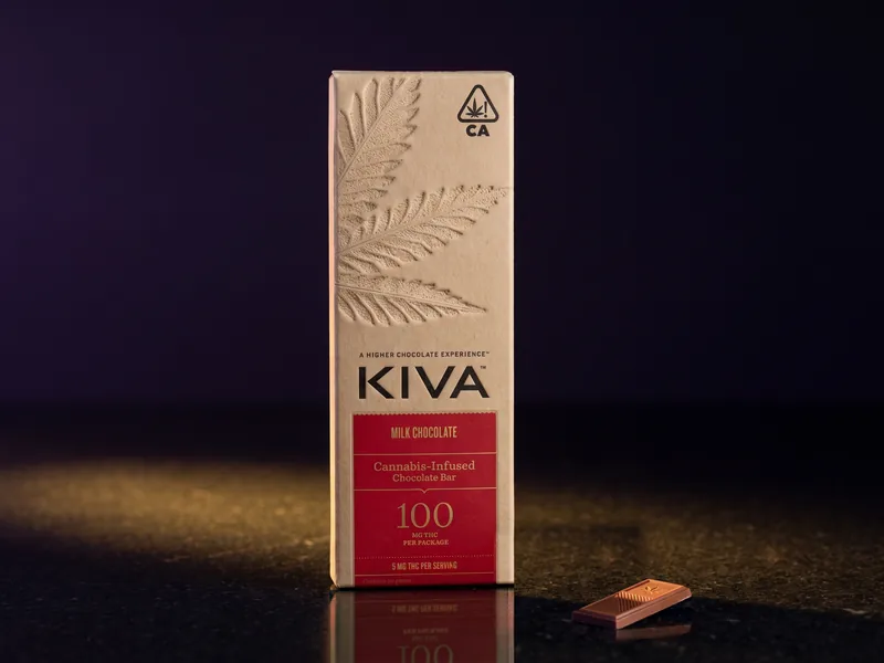 A Kiva Milk Chocolate bar box, kraft with a red panel that reads 100 mg THC per package, and one square of chocolate in front, on black stone.