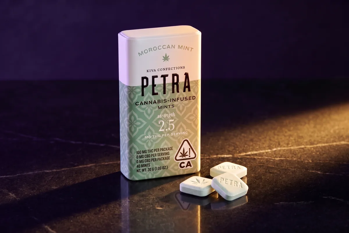 A Kiva Petra Moroccan Mint tin, 2.5 mg THC per serving, with three pale square mints in front, on black stone in gold side light.