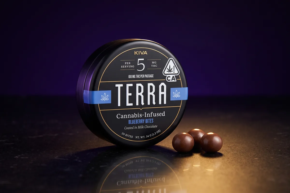 A black Kiva Terra Blueberry Bites tin, 5 mg THC per serving, on its edge next to three milk chocolate bites, on dark stone in gold light.