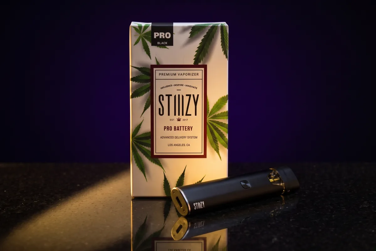 A STIIIZY PRO Black battery box, with one matte black STIIIZY battery lying in front of it, on black stone in gold light.