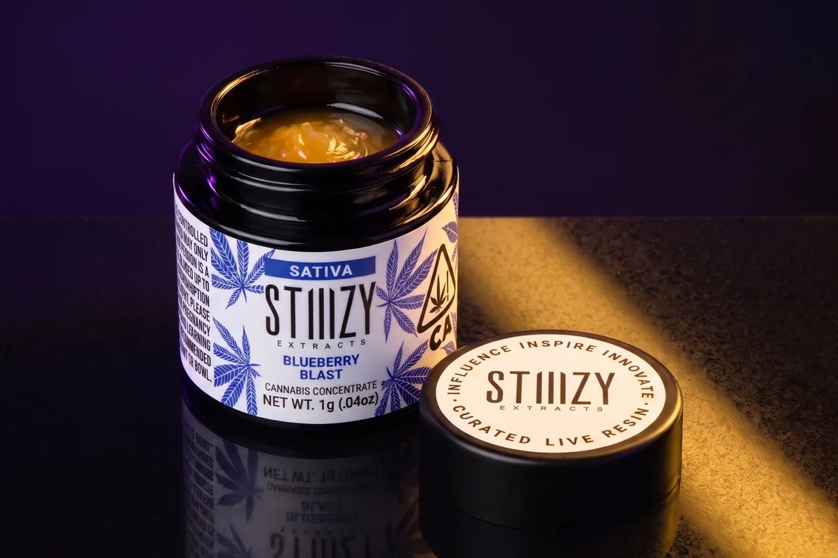 An open STIIIZY Extracts jar of Blueberry Blast sativa sauce, amber inside, with its lid beside it on black stone in gold light.