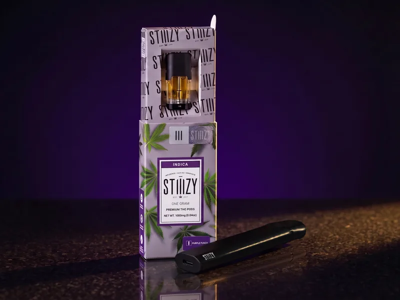 A STIIIZY Purple Punch pod box with its sleeve pulled up to show the pod, and a black STIIIZY PRO battery in front, on black stone in gold light.