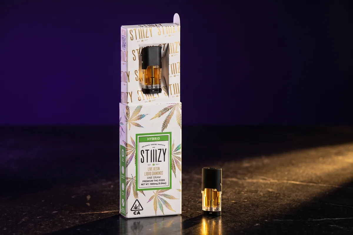 A STIIIZY Live Resin Liquid Diamonds pod box with a green Hybrid label, and one black STIIIZY pod of amber oil beside it, on black stone.