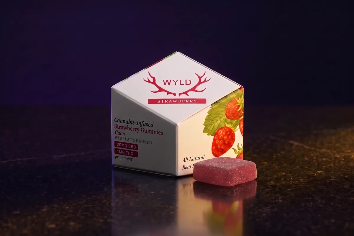 A white WYLD Strawberry gummies box, 20mg CBD and 1mg THC per gummy, with one square ruby gummy in front, on black stone in gold light.