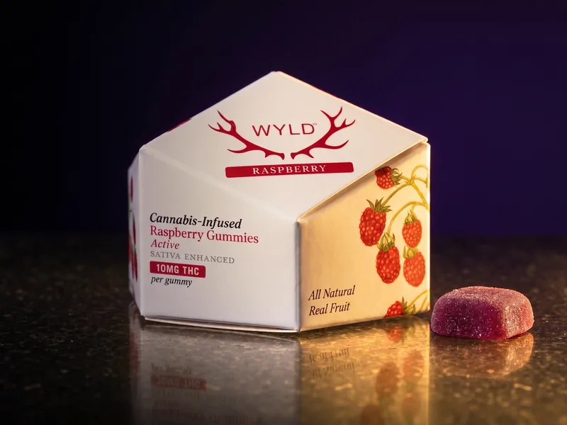 A white WYLD Raspberry gummies carton with red antlers and a red Raspberry band, and one wine-red gummy beside it, on black stone.