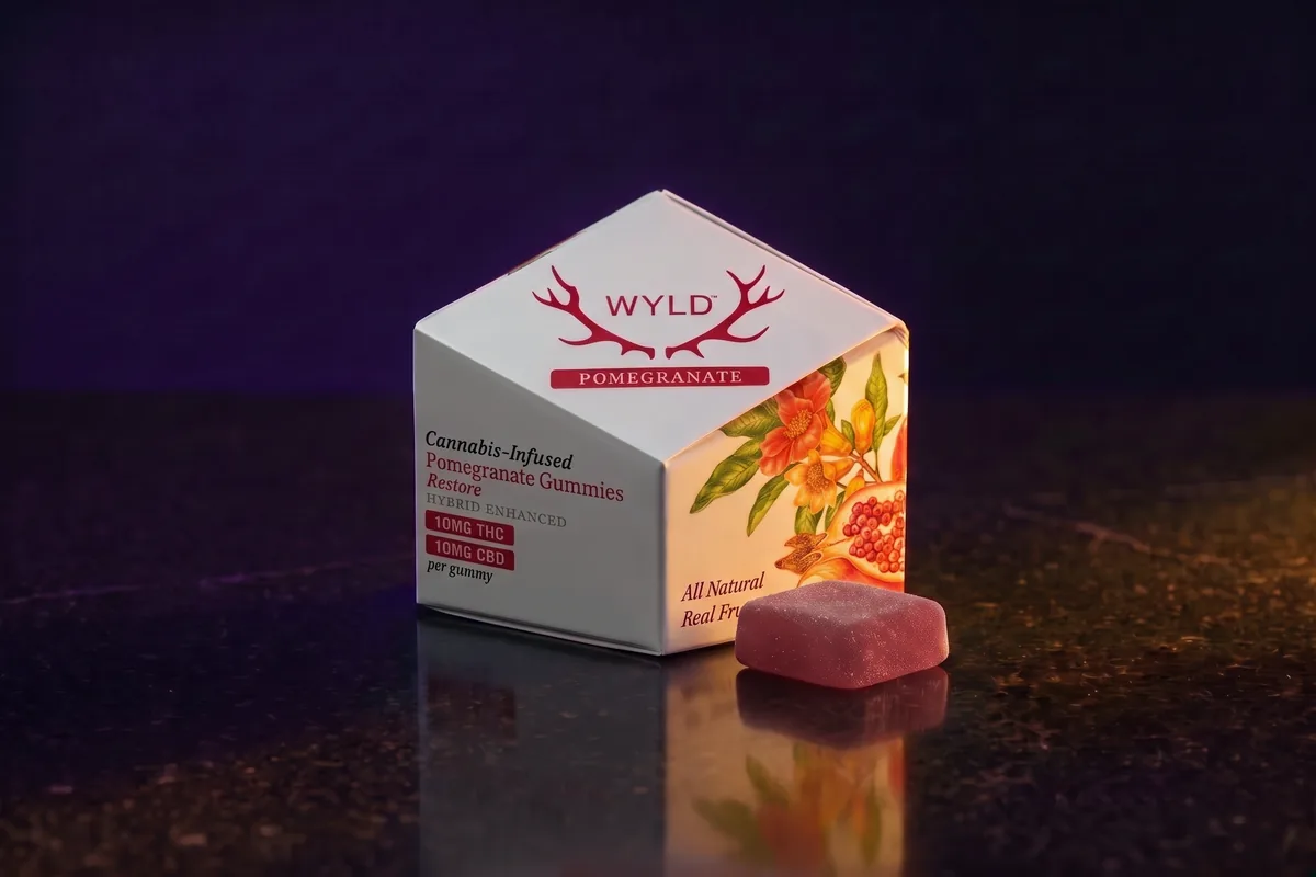 A white WYLD Pomegranate gummies carton, 10mg THC and 10mg CBD per gummy, with one ruby gummy in front of it, on black stone in gold light.