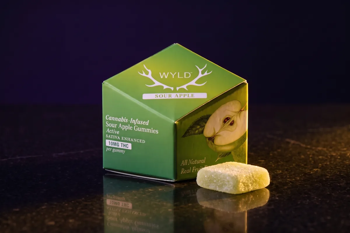 A green WYLD Sour Apple gummies box, 10mg THC per gummy, with one square sugar-coated green gummy in front, on black stone in gold light.